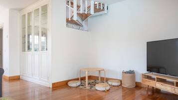 A modern living room with a wooden floor, a television, a white cabinet with glass doors, a small round table, and a staircase.
