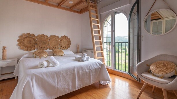 Una camera da letto con soffitto in legno, un letto con lenzuola bianche, uno specchio tondo e vista sull'esterno.