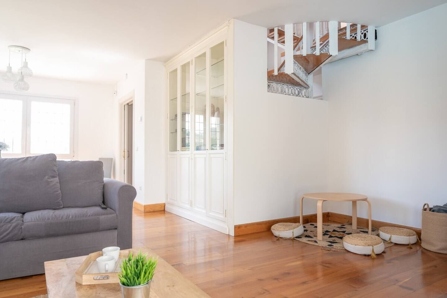 Una sala moderna con un sofá gris, una mesa de centro de madera y una pequeña mesa redonda. Se ve una escalera con barandilla de madera que sube a un nivel superior.