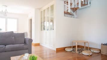 Una sala moderna con un sofá gris, una mesa de centro de madera y una pequeña mesa redonda. Se ve una escalera con barandilla de madera que sube a un nivel superior.
