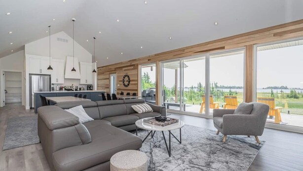 A spacious living area with a large sectional sofa, a round coffee table, and a grey armchair. The room features a modern kitchen in the background, large windows offering a view of the outdoors, and a wooden ceiling. - Huronia Luxury: Lakefront Views and Game Room (Mar)