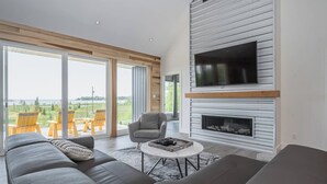 A modern living room with a grey sofa, a grey armchair, and a TV mounted on a white wall. There is a fireplace with a wooden mantel and a large window overlooking a green landscape. - Huronia Luxury: Lakefront Views and Game Room (Mar)