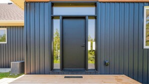 A modern building with a dark exterior, a large sliding glass door, and a wooden deck. - Huronia Luxury: Lakefront Views and Game Room (Mar)