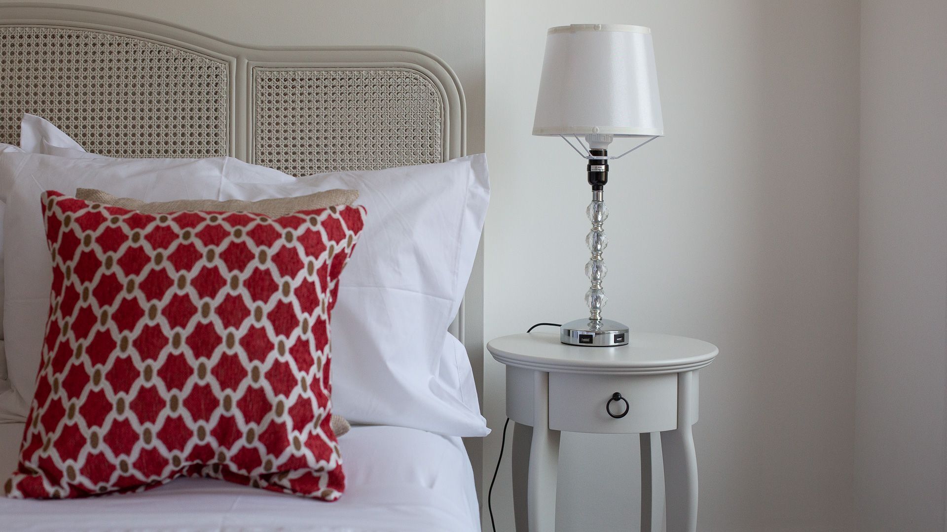 A neatly made bed with white linens and a red patterned pillow, a white bedside table with a lamp, and a wall with a textured headboard.