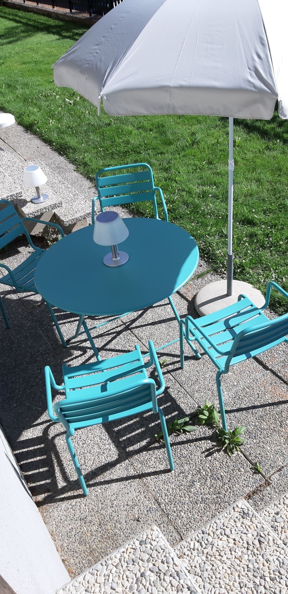 Et rundt bord med en hvid parasol, omgivet af turkise stole, placeret på en grusbelagt terrasse med en græsplæne i baggrunden.