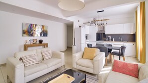 A modern living room with a sofa, armchair, and coffee table. A kitchen is visible in the background with white cabinets and a dining area. - Superb Apartment 100 Meters From the Port of Nice (Nice)