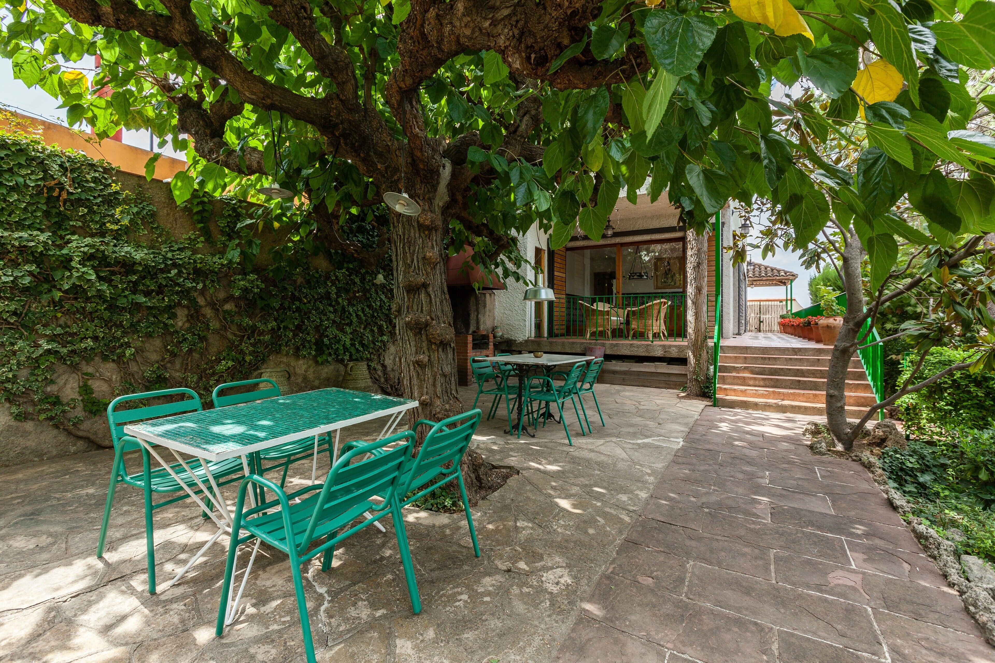 Une terrasse extérieure avec une table et des chaises, un chemin de pierres et un arbre qui offre de l’ombre.