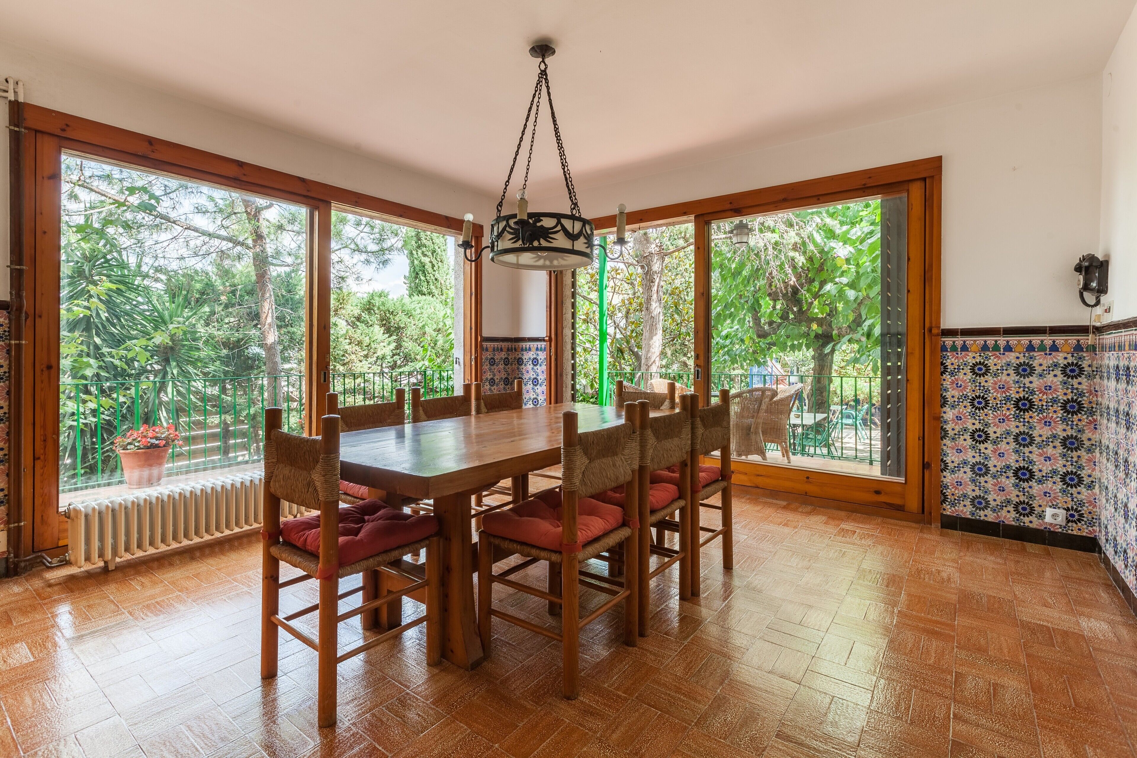 A dining area with a wooden table and chairs, large windows with a view of greenery, and a hanging light fixture.