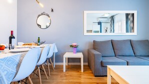 A modern living room with a grey sofa, a dining table set for two, a small white side table with a vase, and a large mirror on the wall. - Holiday Apartment Caracola with Balcony, Covered Terrace & Wi-Fi (Vinaròs)
