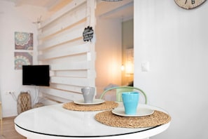Una mesa de vidrio redonda con dos platos y dos tazas, un camino de mesa tejido, una silla verde, un reloj de pared y un televisor de pantalla plana.