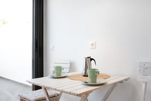 Una mesa pequeña con dos tazas verdes y una cafetera de plata, un mantel tejido y una silla blanca con un cojín a rayas.