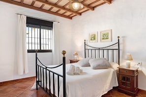 Un dormitorio con cama, mesita de noche, ventana con cortinas y dos cuadros en la pared.