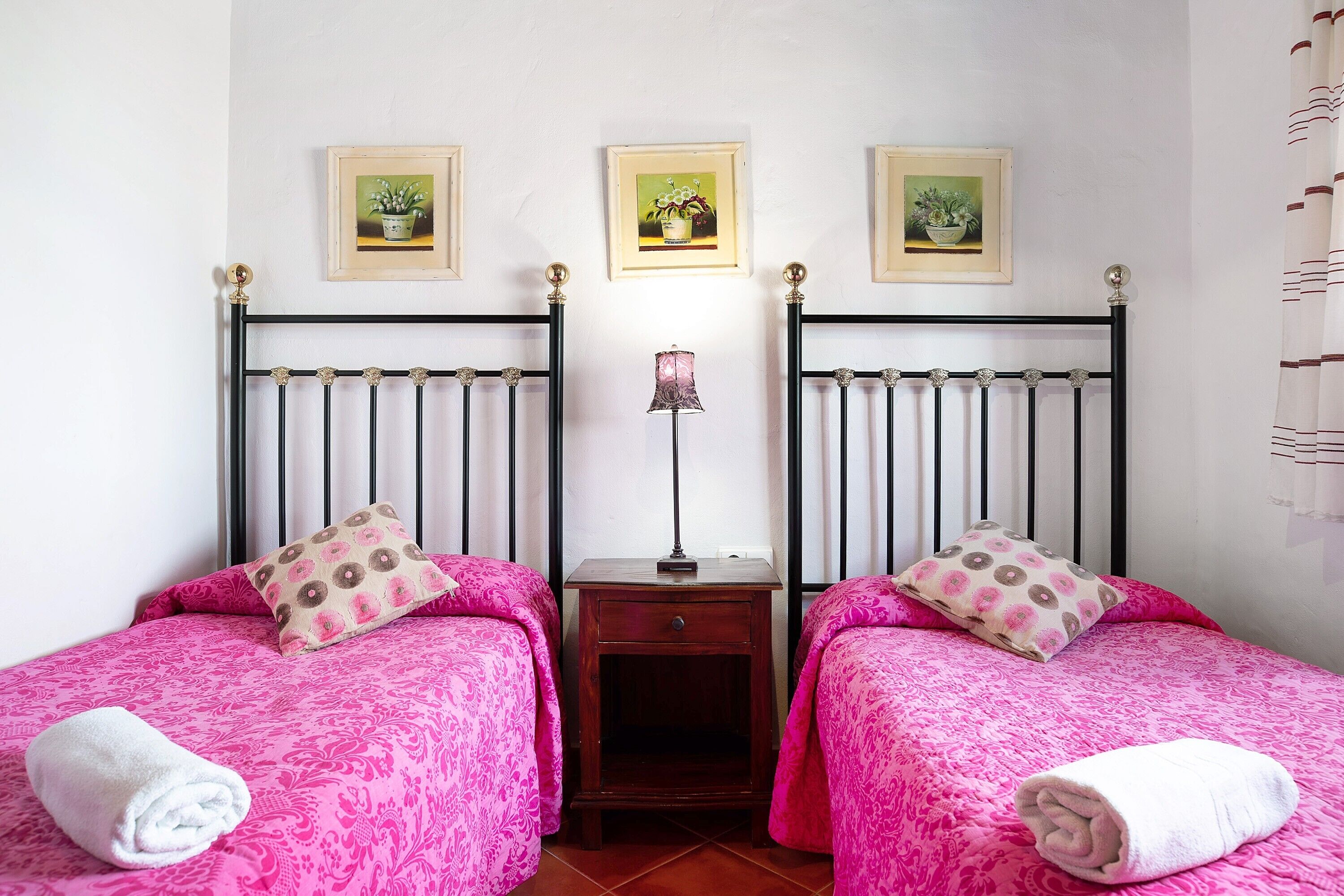 Une chambre avec deux lits, chacun recouvert d’une couvre-lit à motifs roses assorti à des coussins, une petite table de chevet en bois avec une lampe, et trois tableaux encadrés accrochés au mur.