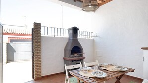 Eine Außenpatio mit einem Backofen aus Ziegeln, weißen Stühlen, einem Holztisch und Blick auf ein Gebäude durch ein Tor.