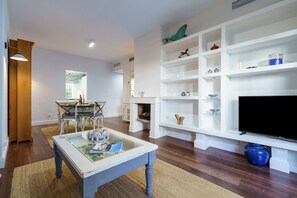 Una sala de estar con una mesa de centro de vidrio, un comedor con mesa y sillas de madera, y un mueble blanco de entretenimiento con estantes.