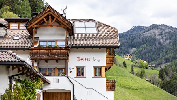A traditional chalet with a wooden facade, a balcony with flowers, and a view of a green hillside. - 'Bolser Apartment Plan de Corones' with Mountain View, Garden & Wi-Fi (Pieve di Marebbe)