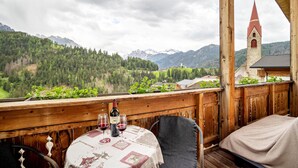 Ein Holzbalkon mit Tisch für Zwei, mit Blick auf eine Berglandschaft und eine Kirche.