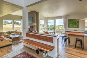 A modern living space with a sofa, coffee table, and a wooden bench. There is a kitchen area with a bar and stools. - Lovely Day | BBQ Grill, Hot Tub, & Ocean Views (Sea Ranch)