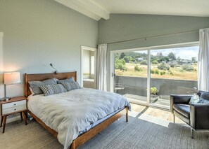 Un dormitorio con una cama grande, una mesita de noche, una silla y vistas al exterior a través de un ventanal corredero.