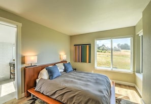 A bedroom with a large bed, two bedside lamps, a window with a view of the outdoors, and a wall-mounted artwork. - Lovely Day | BBQ Grill, Hot Tub, & Ocean Views (Sea Ranch)