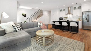 A modern living room with a grey sofa, a round wicker coffee table, and a staircase leading to a kitchen area with white cabinets and a stainless steel refrigerator.