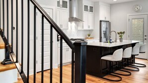 A modern kitchen with white cabinets, a stainless steel range hood, a black island with bar stools, and a wooden staircase with black railings.