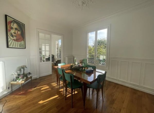 A dining room with a wooden table and chairs, a radiator, and a window with a view of greenery.