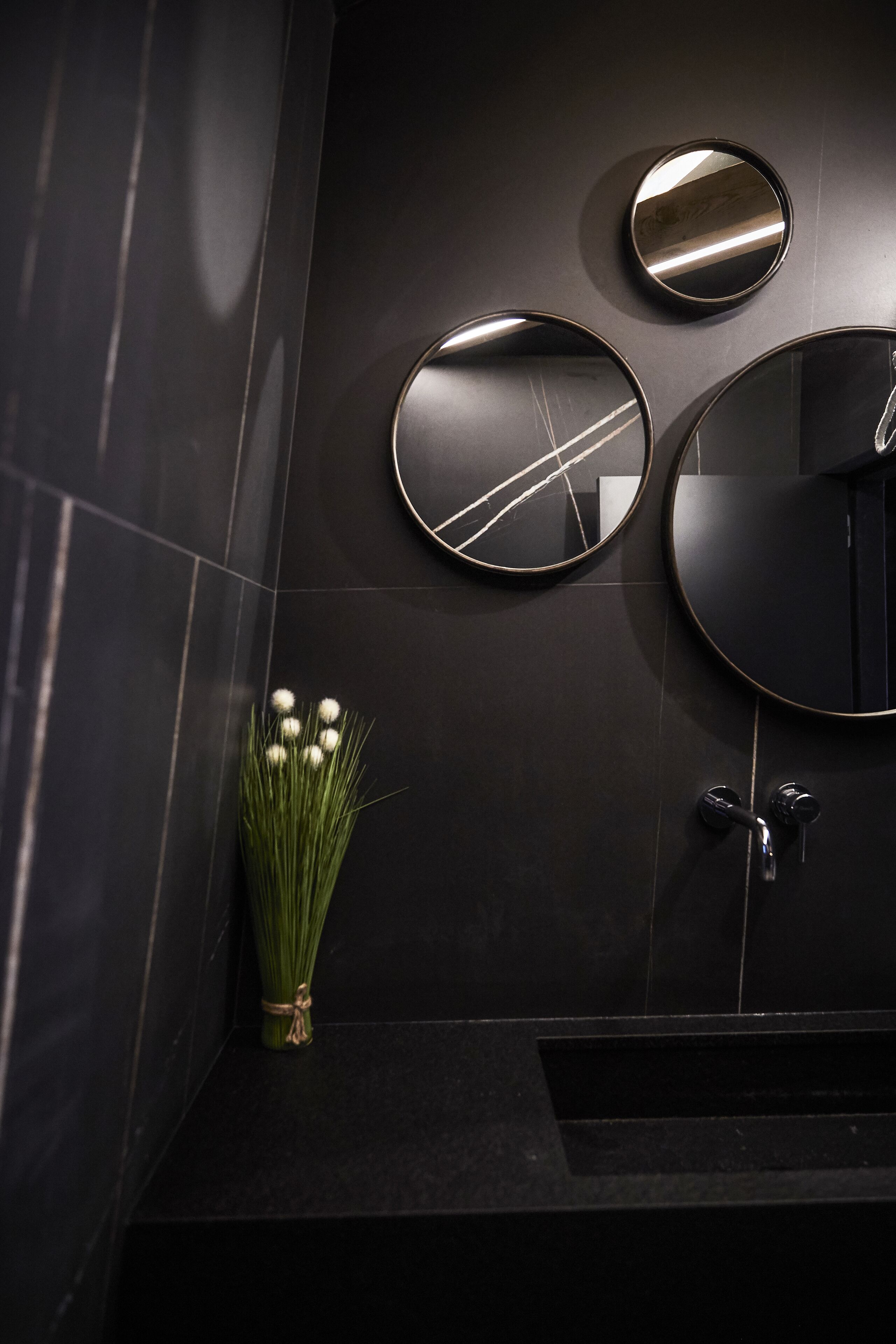 Une salle de bain moderne avec des carreaux de couleur sombre, un miroir rond et un vase rempli de fleurs blanches.
