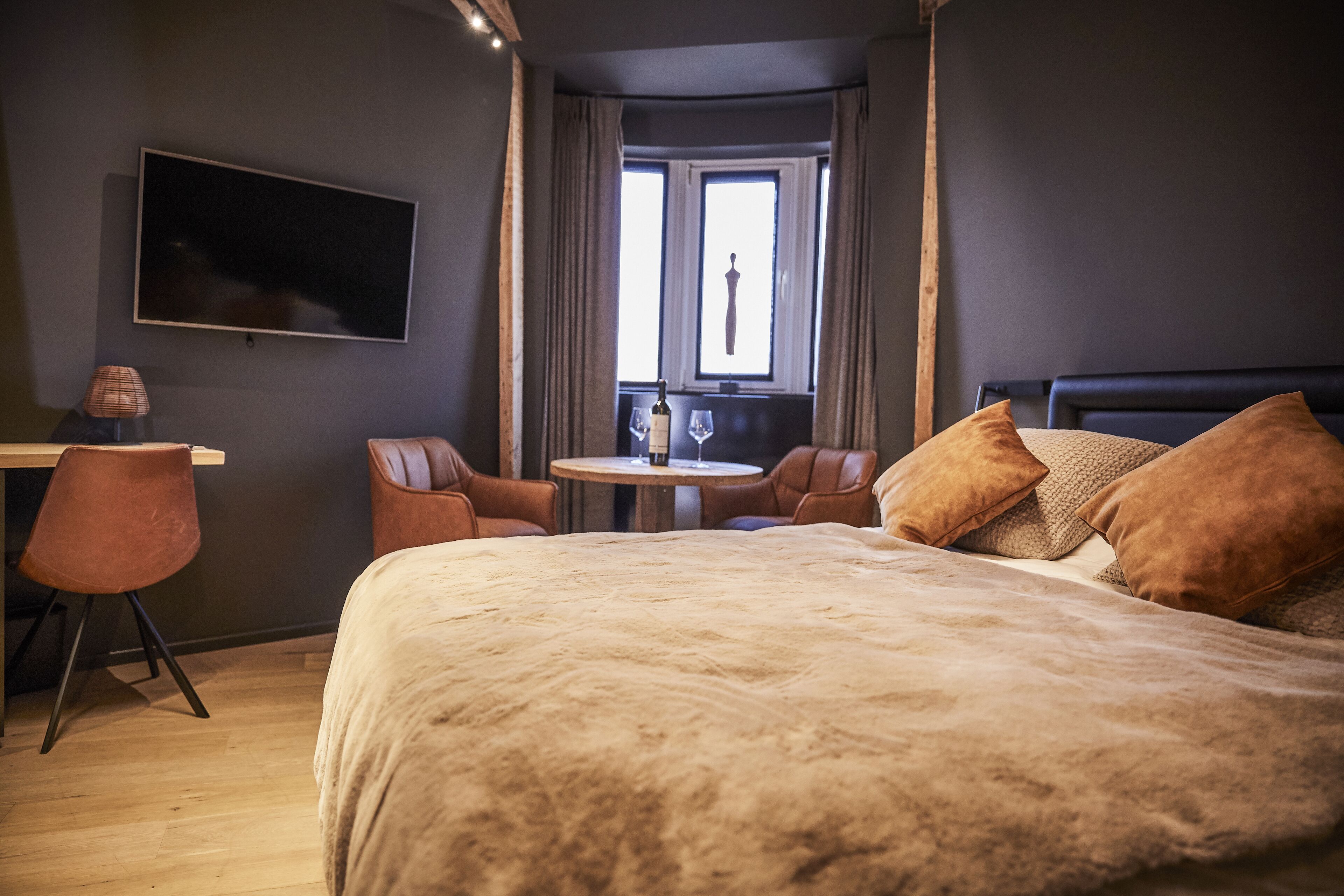 A bedroom with a large bed, a desk, two chairs, a small table, and a TV mounted on the wall.