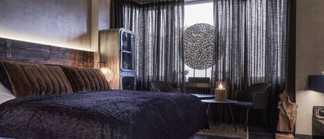 Habitación de hotel moderna con una cama grande, un escritorio y una silla. Se aprecia una ventana con cortinas y un elemento decorativo en la pared.