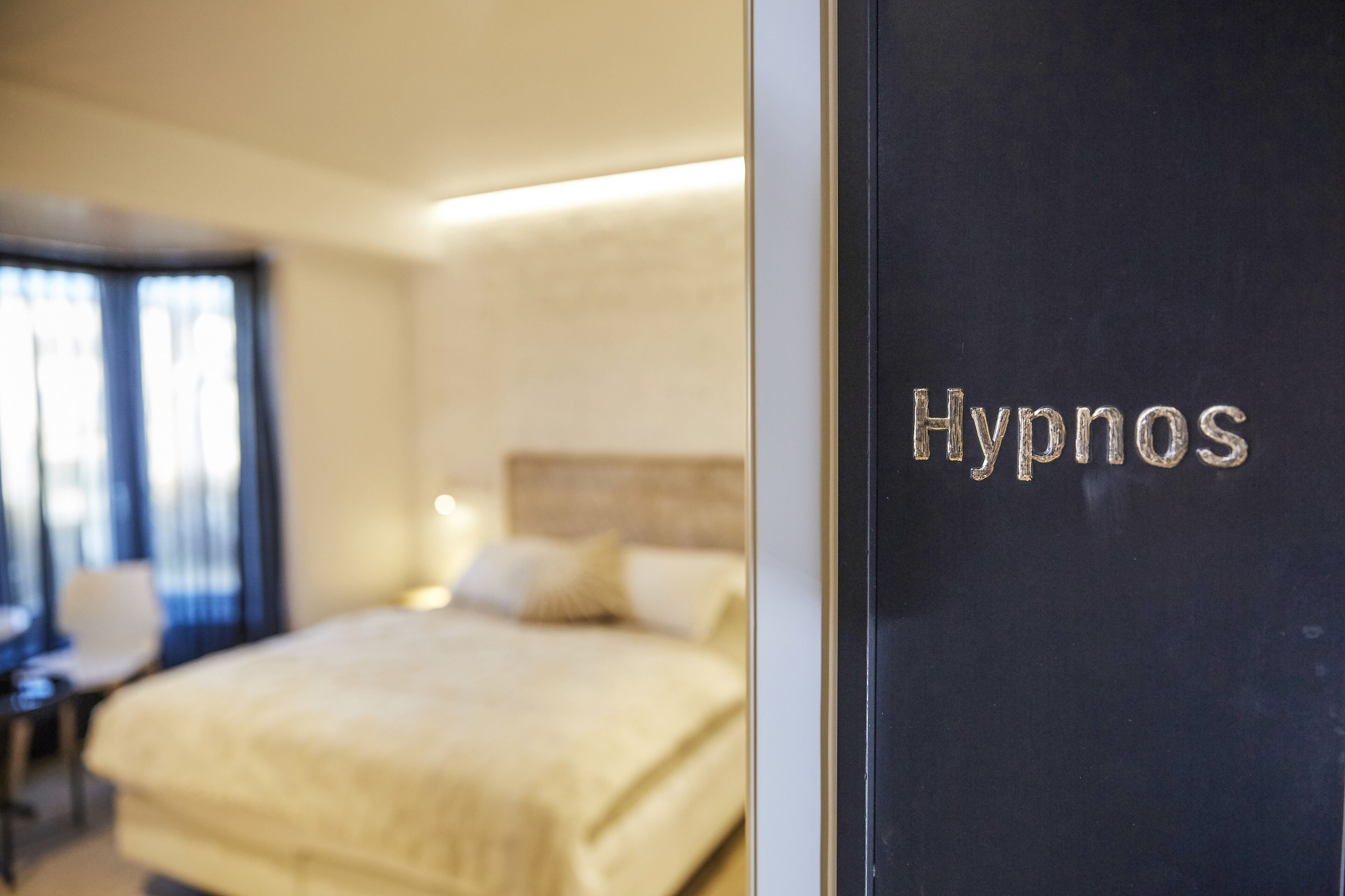 A hotel room with a bed, a chair, and a desk. The room has a sign that says "Hypnos".