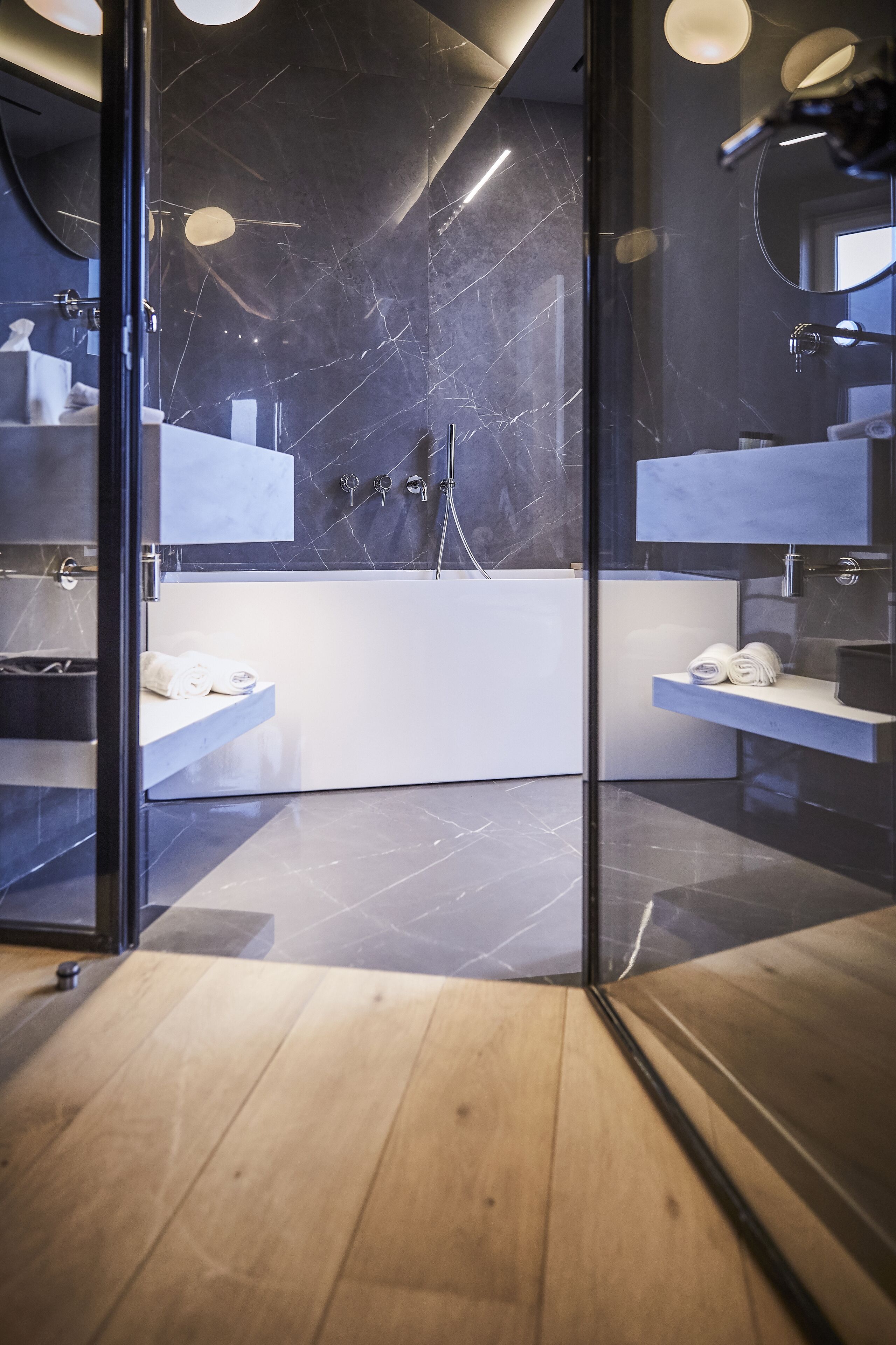 A modern bathroom with a glass partition, marble walls, and wooden flooring.