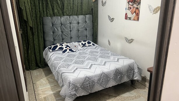 A bedroom with a bed, green curtains, a patterned bedspread, and a wall decoration. - Come to me (Cali)