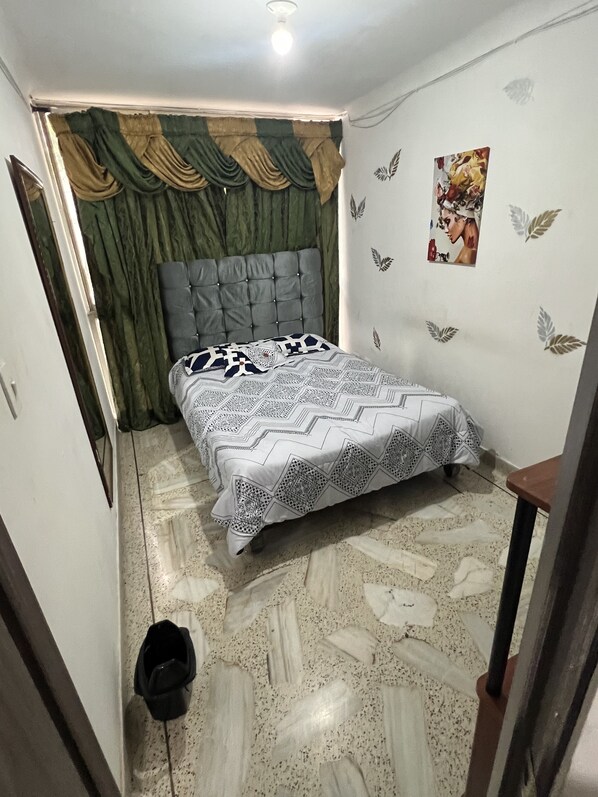 A small, well-lit bedroom with a bed, a headboard, a wall with decorative leaf patterns, and a door with curtains.