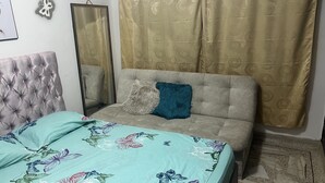 A bedroom with a tufted headboard, a floral bedspread, a grey sofa, a blue pillow, and a patterned curtain. - Come to me (Cali)