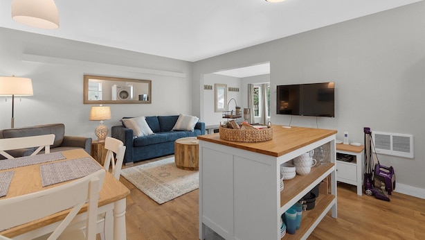 Un salon moderne avec un canapé, une table basse et un téléviseur. Il y a également un coin repas avec une table et des chaises. La pièce est décorée dans des tons neutres et le sol est recouvert de parquet.