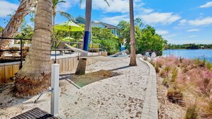 A beachside walkway with hammocks, palm trees, and a view of a house and water.