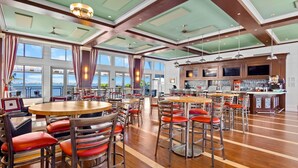 A spacious dining area with multiple round tables and red cushioned chairs, large windows offering a view of the sea, and a bar area with a counter and televisions.