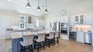Une cuisine spacieuse avec des meubles blancs, un îlot central doté de tabourets, des appareils en acier inoxydable et un éclairage suspendu.