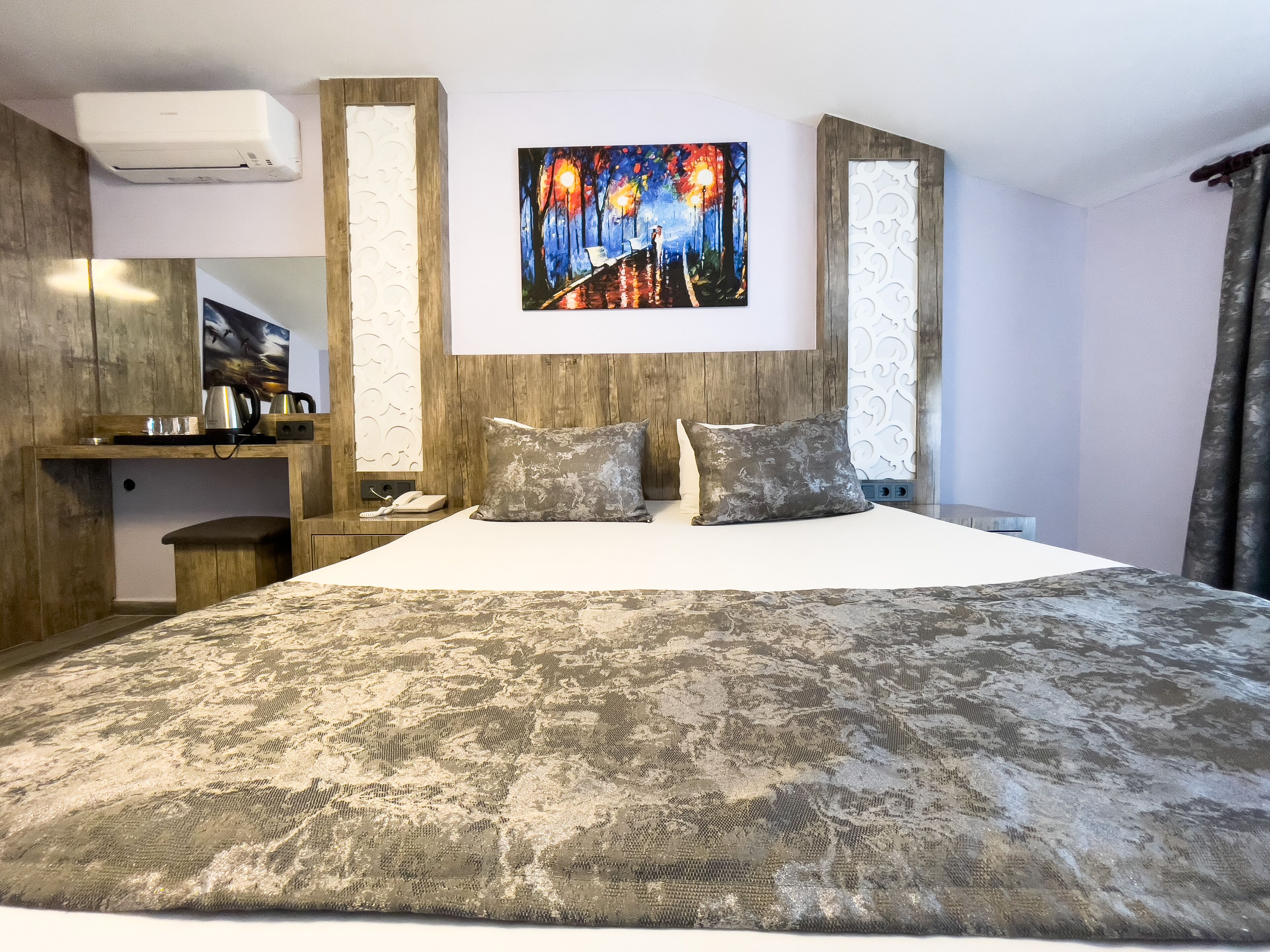 A modern hotel room with a large bed, a painting above the bed, and a wooden headboard.