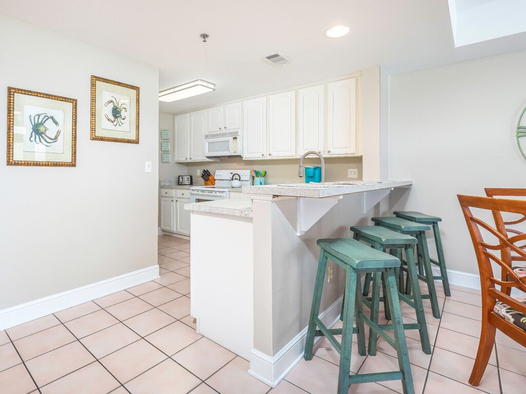 A kitchen with white cabinets, a microwave, and a sink. There are green stools and a wooden chair.