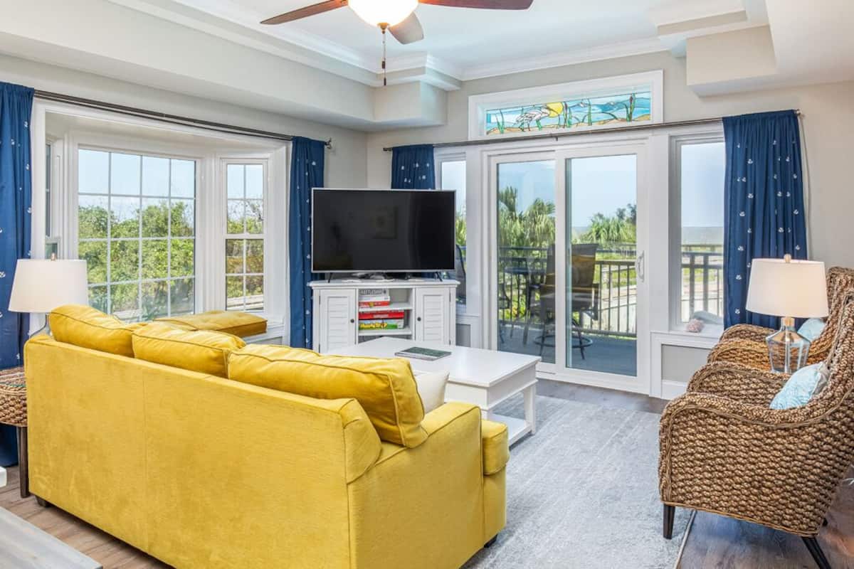 A living room with a yellow sofa, a wicker chair, a flat-screen TV, and a balcony with a view.