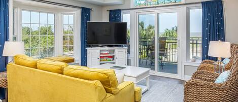 A living room with a yellow sofa, a wicker chair, a flat-screen TV, and a balcony with a view.