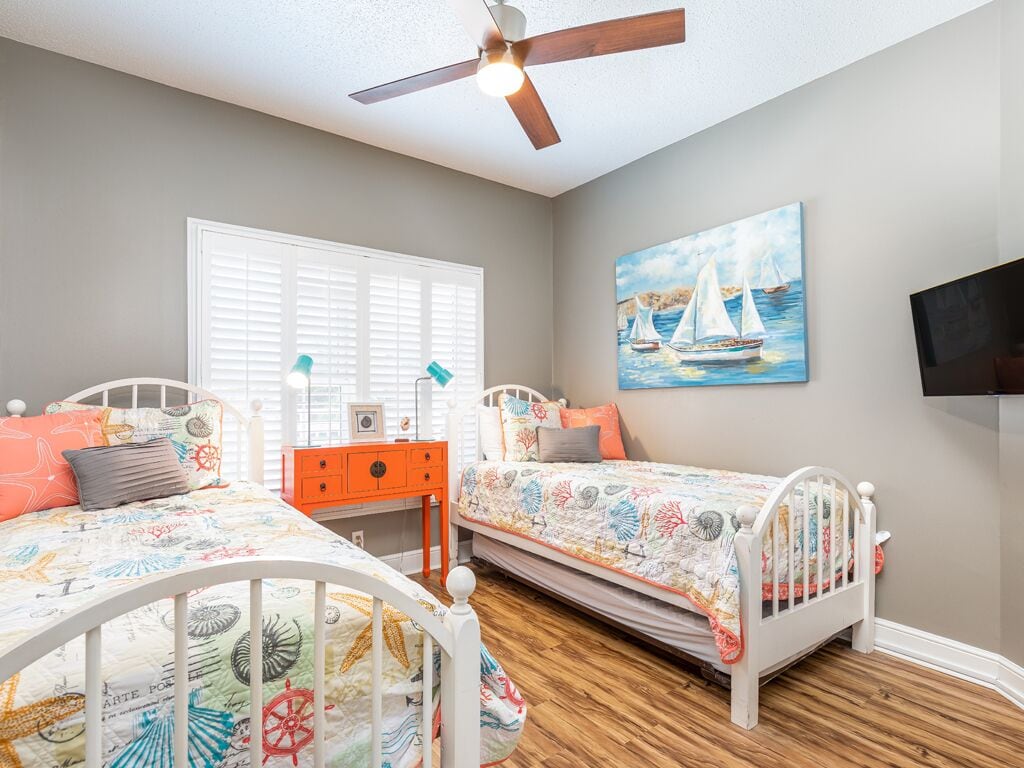 A bedroom with two beds, a ceiling fan, a painting of sailboats, and a TV on the wall.