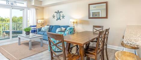 Un coin repas avec une table et des chaises en bois, un ventilateur de plafond et une vue sur la plage, accessible par une porte coulissante.