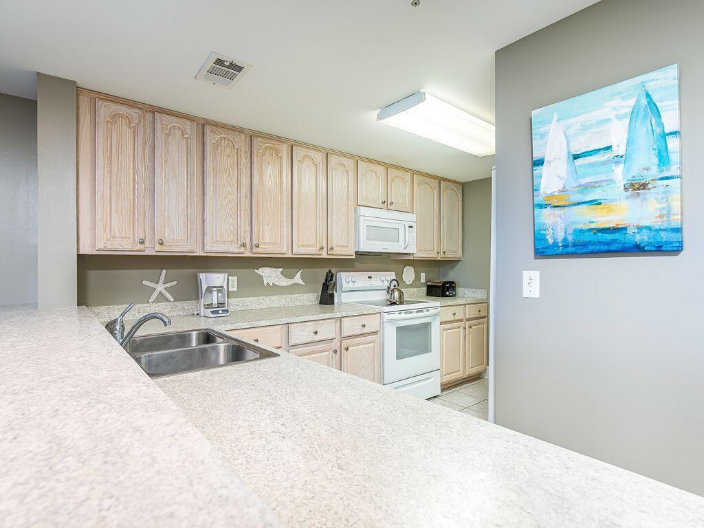 A kitchen with light wooden cabinets, a white microwave, a stove, and a sink. There is a painting of sailboats on the wall.