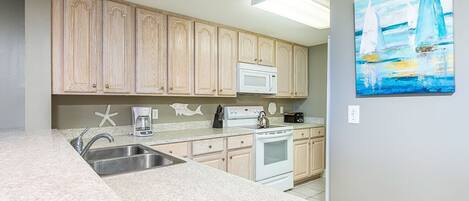 A kitchen with light wooden cabinets, a white microwave, a stove, and a sink. There is a painting of sailboats on the wall.