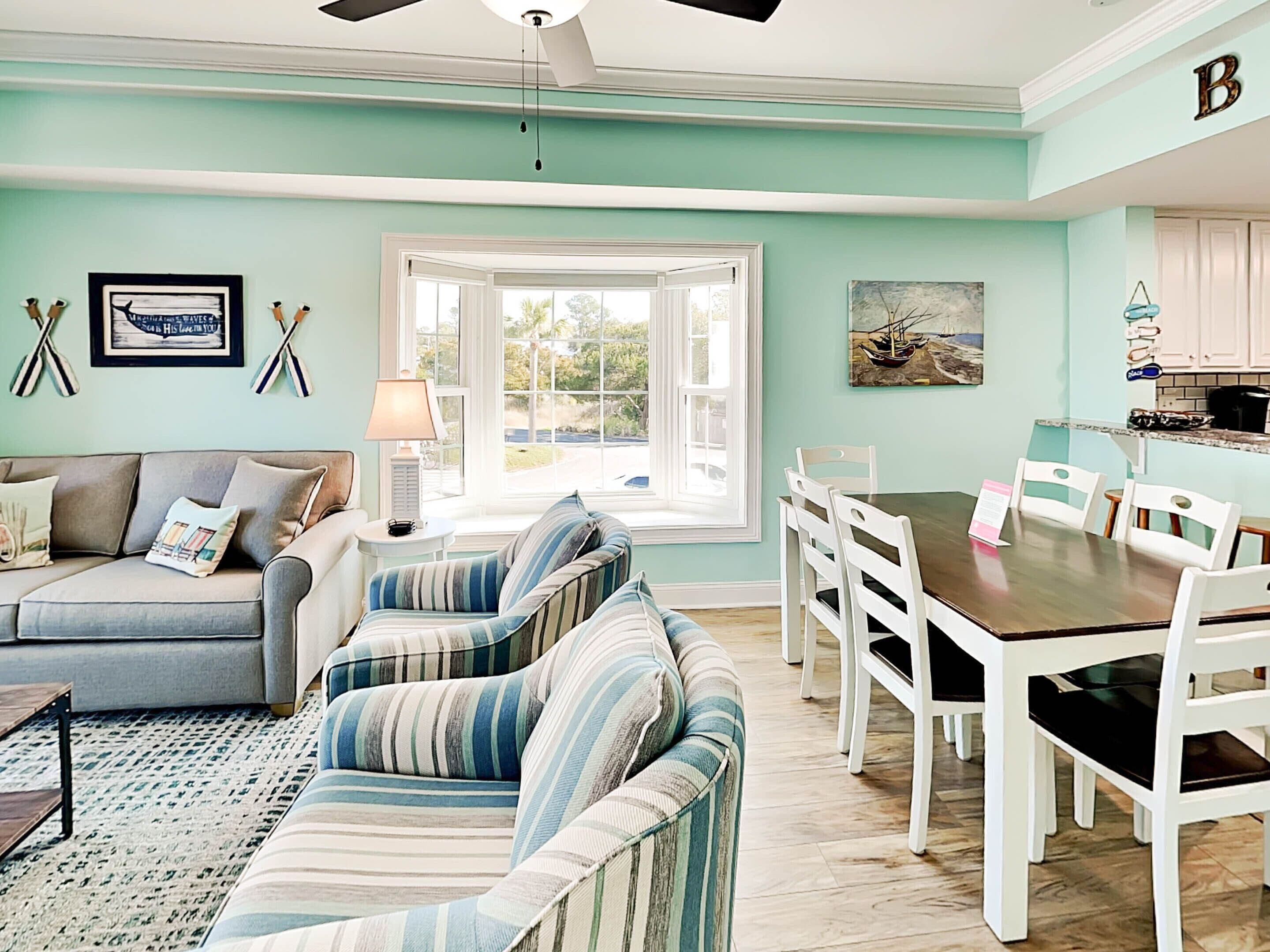 A living room with a sofa, striped armchairs, a dining table with chairs, a ceiling fan, and a window with a view of greenery.
