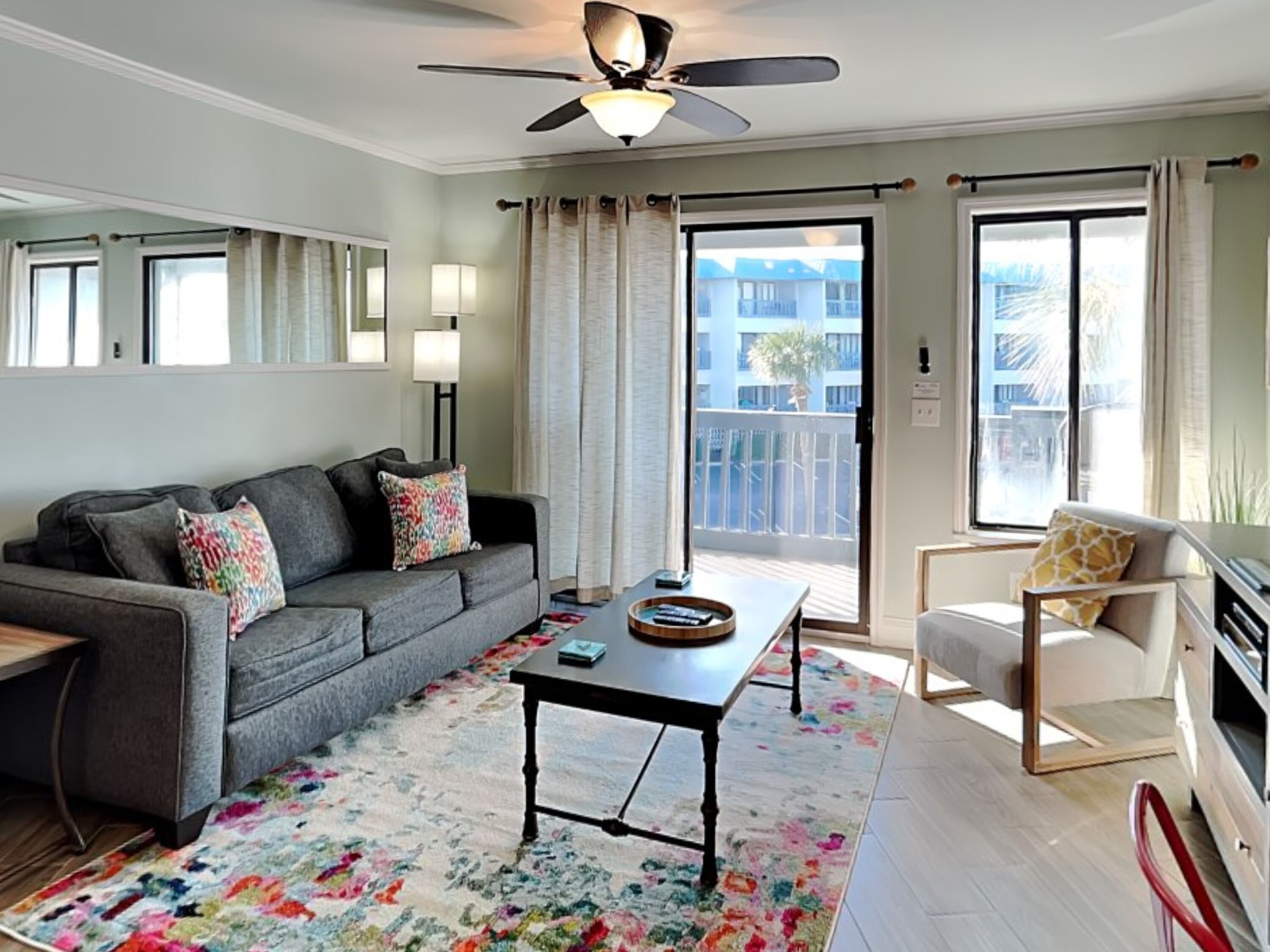 A living room with a gray sofa, a coffee table, a floral rug, a ceiling fan, and a view of a balcony with a railing.