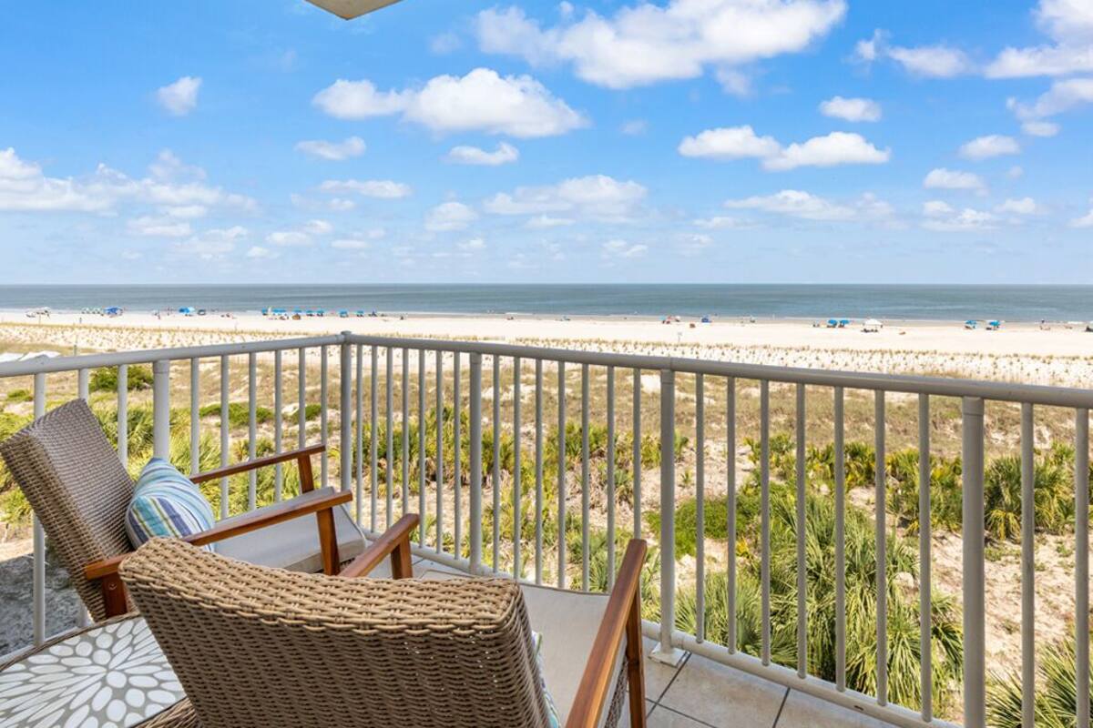 A balcony with wicker chairs and a table, overlooking a beach with palm trees and a clear sky.